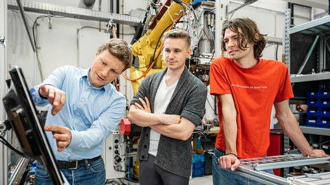 Photo of two students and their lecturer. The lecturer is showing something to his students on a screen.