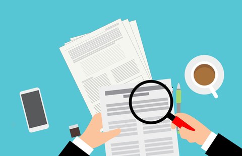 The graphic shows in the foreground a sheet of paper with text held by one hand, over which a magnifying glass is held with the other hand. In the background, from left to right: smartphone, USB stick, stack of paper with text, pen, coffee cup.