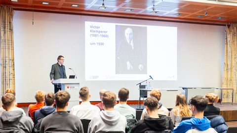 Blick in einen Vortragssaal. Ein Mann steht an einem Rednerpult. Im Vordergrund sieht man Schüler:innen, die zuhören.