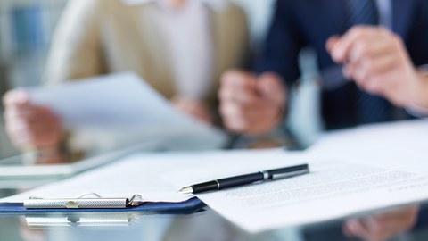 Photo of documents on a table. In the background, two people can be seen looking at a document held by another person. 
