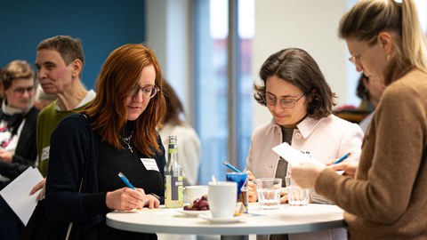 Several women stand at a bar table at the New Year's reception of the Women@DDc network, writing notes and chatting over drinks in a relaxed atmosphere.