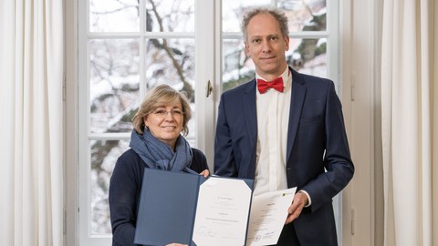 Zwei Personen stehen vor einem Fenster und halten geöffnete Mappen mit Urkunden; winterliche Landschaft im Hintergrund.