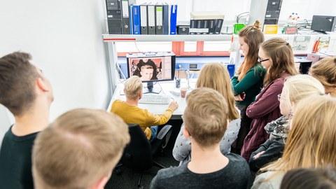 The photo shows a group of young people in an office. They are standing in a semicircle around a workstation with a monitor and keyboard. A man is sitting at this place. Their eyes are fixed on the monitor.
