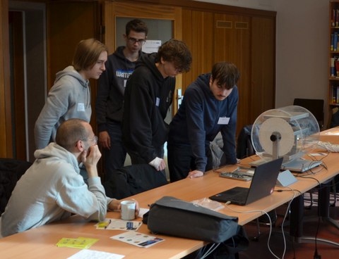 Mehrere Jugendliche beugen sich über einen Tisch und arbeiten gemeinsam an einem technischen Experiment mit Laptop und Detektor-Aufbau.