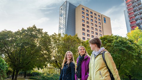 The photo shows three female students. They are walking together through a park and talking to each other. Student residences can be seen in the background.