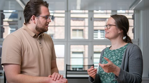 Eine Frau und ein Mann stehen zueinander gewandt an einem Tisch und unterhalten sich. Es handelt sich um eine Studienberaterin und einen Klienten.