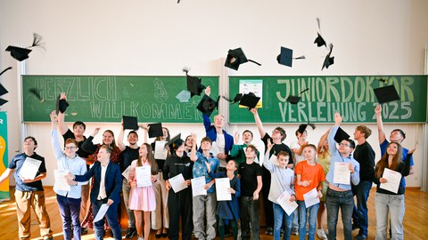 Ca. 25 Kinder und Jugendliche stehen in 2 Reihen vor einer grünen Tafel und werfen Doktorhüte hoch. Auf der Tafel steht "Herzlich willkommen. Juniordoktor-Verleihung 2025."
