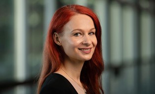 Portrait of a woman with long red hair smiling warmly. She is wearing a necklace and a black top.