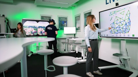 Photo: A person with blonde hair points at a screen displaying a geometric diagram. In the background, another person wears a VR headset and a second screen is visible.