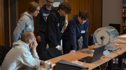 Mehrere Jugendliche beugen sich über einen Tisch und arbeiten gemeinsam an einem technischen Experiment mit Laptop und Detektor-Aufbau.
