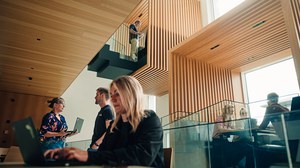 Several people are working in a modern, multi-story atrium featuring wooden slats and glass railings. A young woman in the foreground is typing on a laptop, while others are conversing, standing on an upper level, or sitting at tables.
