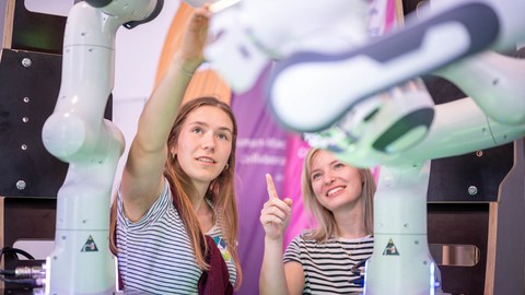 Foto von zwei jungen Frauen in schwarz-weiß gestreiften T-Shirts, die inmitten von weißen Roboterarmen stehen und diese betrachten.