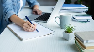 Photo of a person sitting at a desk. The person is writing something on a notepad and at the same time has their other hand on the keyboard of a notebook.