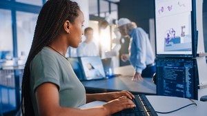 A focused young woman with braids and a green T-shirt types on a keyboard. She looks at two monitors displaying computer code and a 3D model of a robotic hand. In the background, two other people interact.