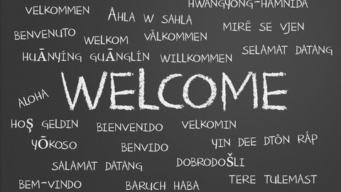 Die Darstellung zeigt eine schwarze Tafel. An diese wurde mit weißer Kreide groß das Wort "Welcome" in die Mitte geschrieben. Drum herum stehen viele kleinere Begriffe. Sie sind die Übersetzung dieses Wortes in verschiedenen Sprachen.