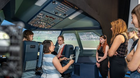 Foto von mehreren Schülerinnen, die sich im Flugsimulator der TU Dresden aufhalten. Eine Frau kniet zwischen den Sitzen von Pilotin und Co-Pilotin und spricht zu der Gruppe der Teilnehmenden