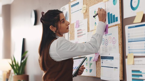 Foto einer Person an einer Pinnwand mit vielen Zetteln, auf denen Diagramme zu sehen sind. 