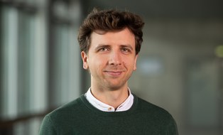 Ein Mann mit kurzen braunen Haaren lächelt in die Kamera. Er trägt einen grünen Pullover und darunter ein Hemd. 