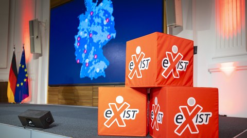 drei Sitzwürfel mit der Aufschrift "exist" stehen zu einer Pyramide gestapelt vor einer Leinwand, auf der eine Deutschlandkarte abgebildet ist. auf der Deutschlandkarte sind zehn Städte markiert - die Gewinner des StartUp Factory Wettbewerbs.