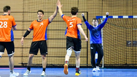Wir sehen 4 Männer in sportlichen Outfits vor einem Handballtor in einer Halle, die beiden in der Mitte klatschen sich ab. Der Mann im Tor hebt seine Arme und ballt die Fäuste.