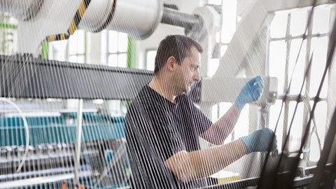 Man sieht die Fäden einer Webmachschine. Im Hintergrund arbeitet ein Mann.