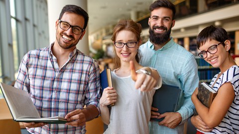 Picture of four students in a library, smiling happily and looking at the camera with books in their hands.