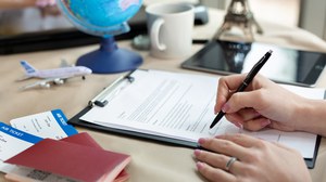 Photo of a person who is filling out a form with a pen. Next to it are passports and in the background is a globe and a small model airplane.