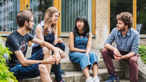 Four students sit on one staircase. On the outside there are two men, in the middle two women. The students talk to each other. 