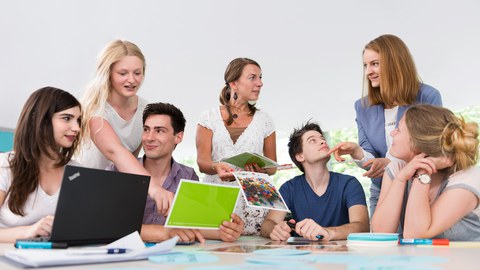 This photo shows several people during a workshop. Five students are sitting at a large table on which various office utensils are lying. Two women stand at the table and turn to the students.