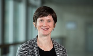 Eine Frau mit kurzen braunen Haaren lächelt in die Kamera. Sie trägt einen schwarz-weiß-karierten Blazer. 