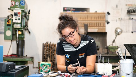 A student is sitting at a workbench. On the workbench and in the background you can see many different tools. The student is looking concentrated on the tool she is holding in her hand. 