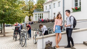 Two students stand on the stairs in front of the cafeteria and talk. Other students are standing in the background. 