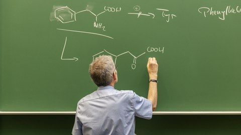 A lecturer writes chemical formulas on a green chalkboard during a lecture.