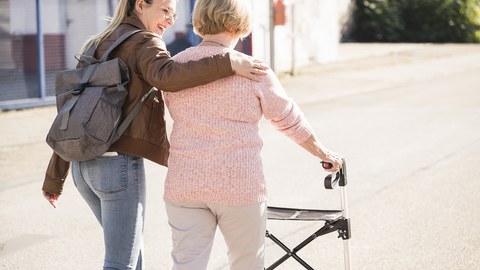 Foto von zwei Personen, die miteinander spazieren gehen. Die ältere Person hat eine Gehhilfe.