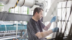 Man sieht die Fäden einer Webmachschine. Im Hintergrund arbeitet ein Mann.