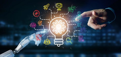 Foto einer menschlichen Hand im oberen rechten Bildrand und einer Roboterhand im unteren linken Bildrand. Beide Händen zeigen mit dem Zeigefinger auf eine visualisierte Darstellung einer Glühbirne mit verschiedenen Icons, die um diese herum angeordnet sind. 
