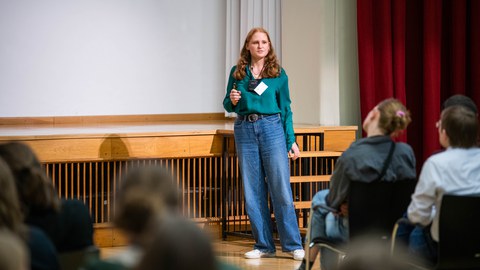Eine Studentin hält einen Vortrag in einem Saal vor mehreren Zuschauerinnen und Zuschauern.