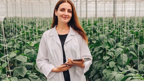 A woman in a white lab coat stands in a greenhouse filled with green plants. She holds a tablet in her hands and smiles, indicating research or work in the field of agriculture or botany.