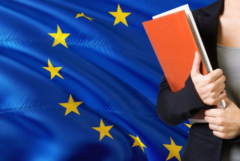  Young woman standing with the European Union flag in the background. Female holding books, orange blank book cover. 
