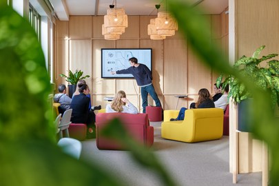 An open, modern seminar room with wooden walls and large windows allowing natural light to enter. A lecturer in dark clothing stands in front of a screen displaying scientific diagrams and points to a graphic. The participants, a diverse group of men and women, are seated comfortably in colorful armchairs—yellow and red—listening attentively. The atmosphere is informal, with woven pendant lights and plenty of green plants, giving the room a vibrant and inviting feel.