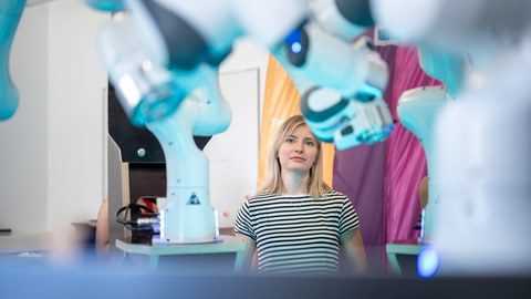 Zwischen Roboterarmen steht ein Mädchen im schwarz-weiß gestreiften T-Shirt und betrachtet diese.