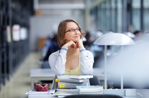 Das Foto zeigt eine Studentin in der Bibliothek. Sie sitzt einen einem Tisch, auf welchem ein Bücherstapel steht. Mit ihren Ellenbogen steht sie auf diesem Bücherstapel und stützt mit ihren Händen ihren Kopf.
