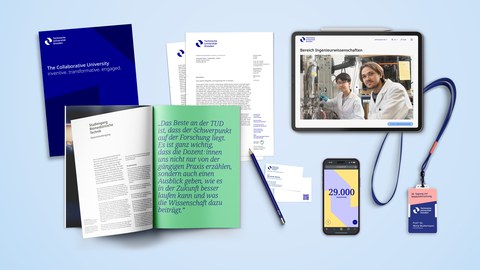 Various branding elements of TU Dresden including a dark blue booklet with white text, an open magazine with green and white text, a letterhead, a pen, a smartphone displaying '29,000', a tablet showing a webpage, and an ID badge with a blue lanyard.