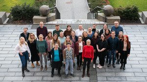 Group photo of a large group of people standing outside on stairs. They are looking at the camera and smiling. In the background, a paved path, green spaces, and bushes can be seen.