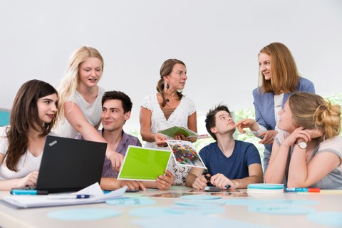 Auf diesem Foto sind mehrere Personen während eines Workshops zu sehen. Fünf Studierende sitzen an einem großen Tisch, auf welchem unterschiedliche Büroutensilien liegen. Zwei Frauen stehen am Tisch und wenden sich den Studierenden zu. 