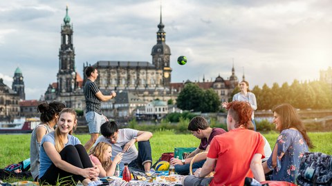 Foto: Gruppe von Studenten an der Elbe. Sie spielen Volleyball und sitzen plaudernd auf der Wiese.
