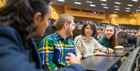 4 Personen im Hörsaal im Vordergrund, die mit einander sprechen, im Hintergrund Blick ins Auditorium des Hörsaals