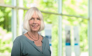 Beate Herm ist als Porträt bis zur Brust abgebildet. Sie trägt weißes Haar als Bob geschnitten, eine helle petrolfarbene Strickjacke und eine Kete mit Glassteinen. Sie lächelt.