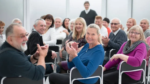 Foto von älteren Menschen in einer Seminarveranstaltung, sie sitzen und klatschen in die Hände. Zwei Frauen schauen freundlich in die Kamera.