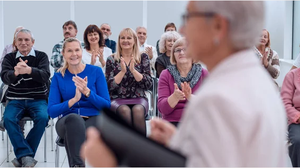 Foto von älteren Menschen in einer Seminarveranstaltung, im Vordergrund eine ältere Dame, die anderen Menschen klatschen